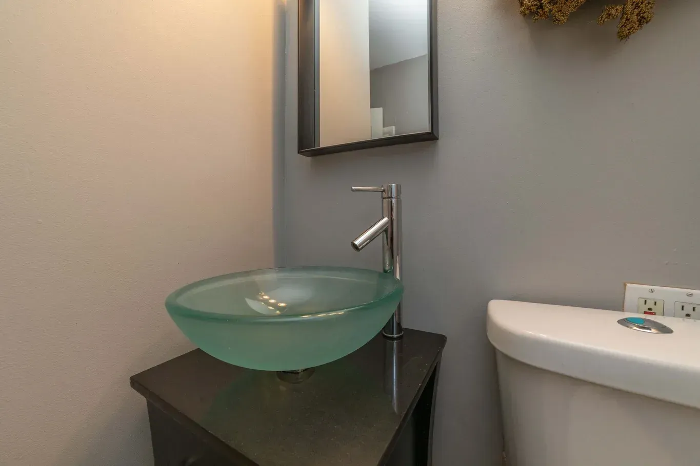 Small bathroom with glass vessel sink on a dark cabinet, mirror, and toilet. Gray walls.