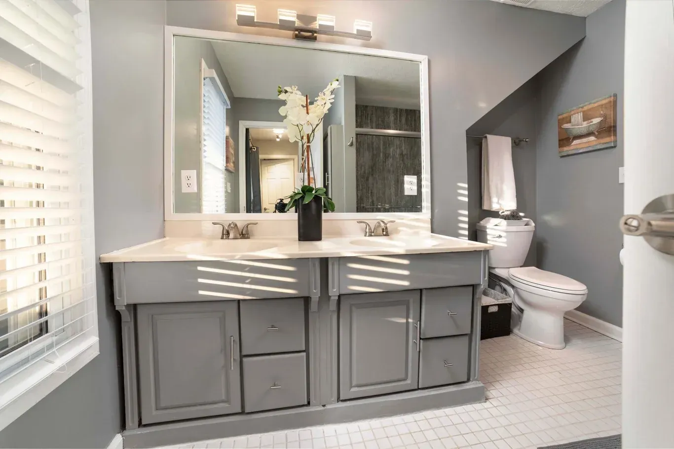 Grey bathroom with double vanity, toilet, and a window with blinds.