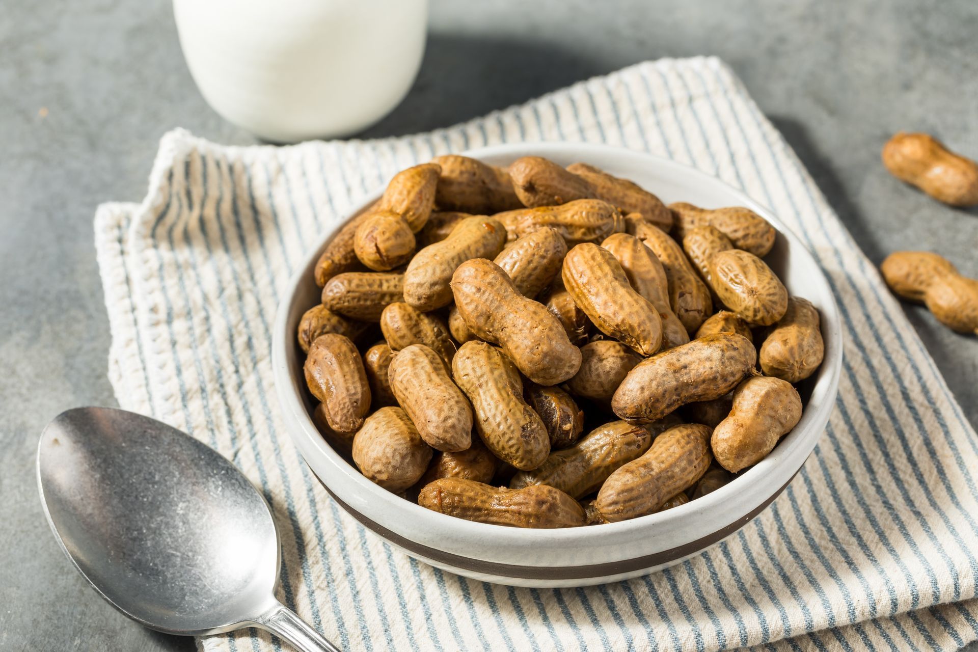Bowl of boiled peanuts on a striped napkin, with a spoon and a glass in the background.