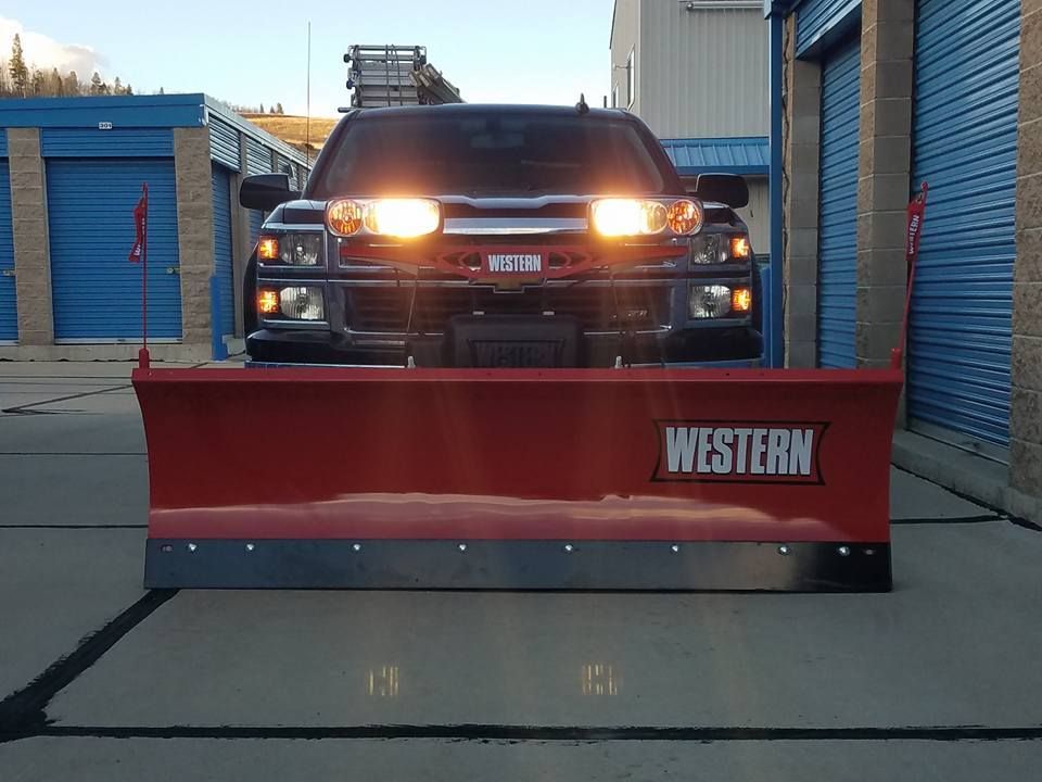 Truck with a red Western snowplow. Headlights on. Parked in front of storage units.