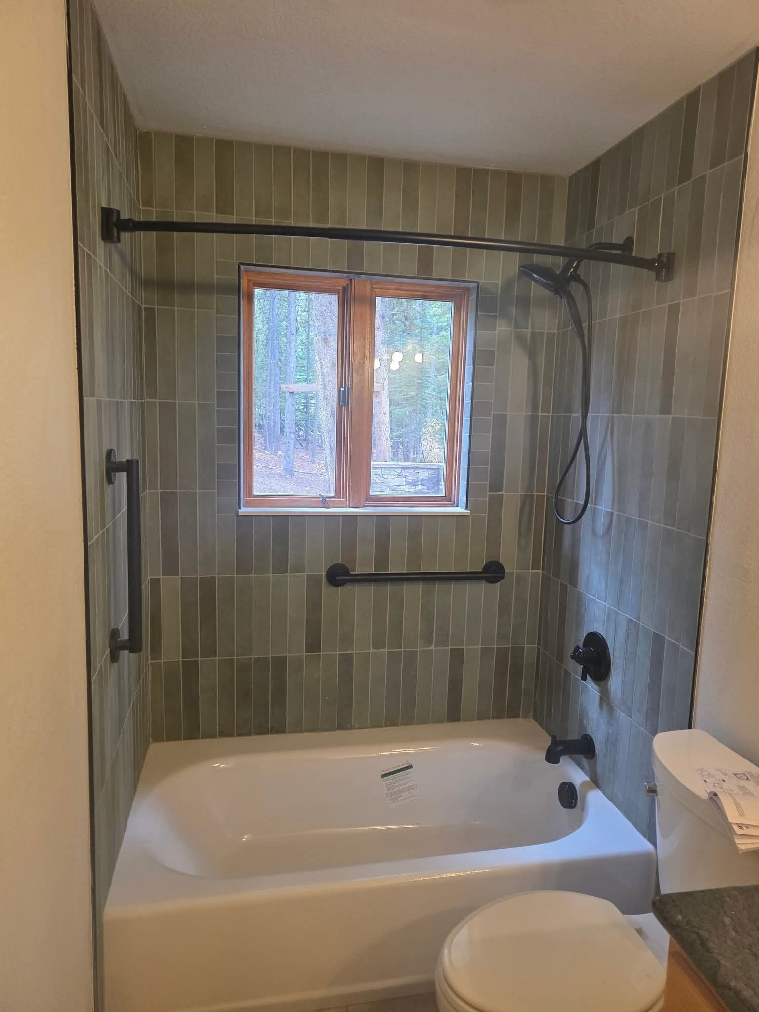 Bathroom with green tiled walls, tub, window, grab bars, and shower fixtures.