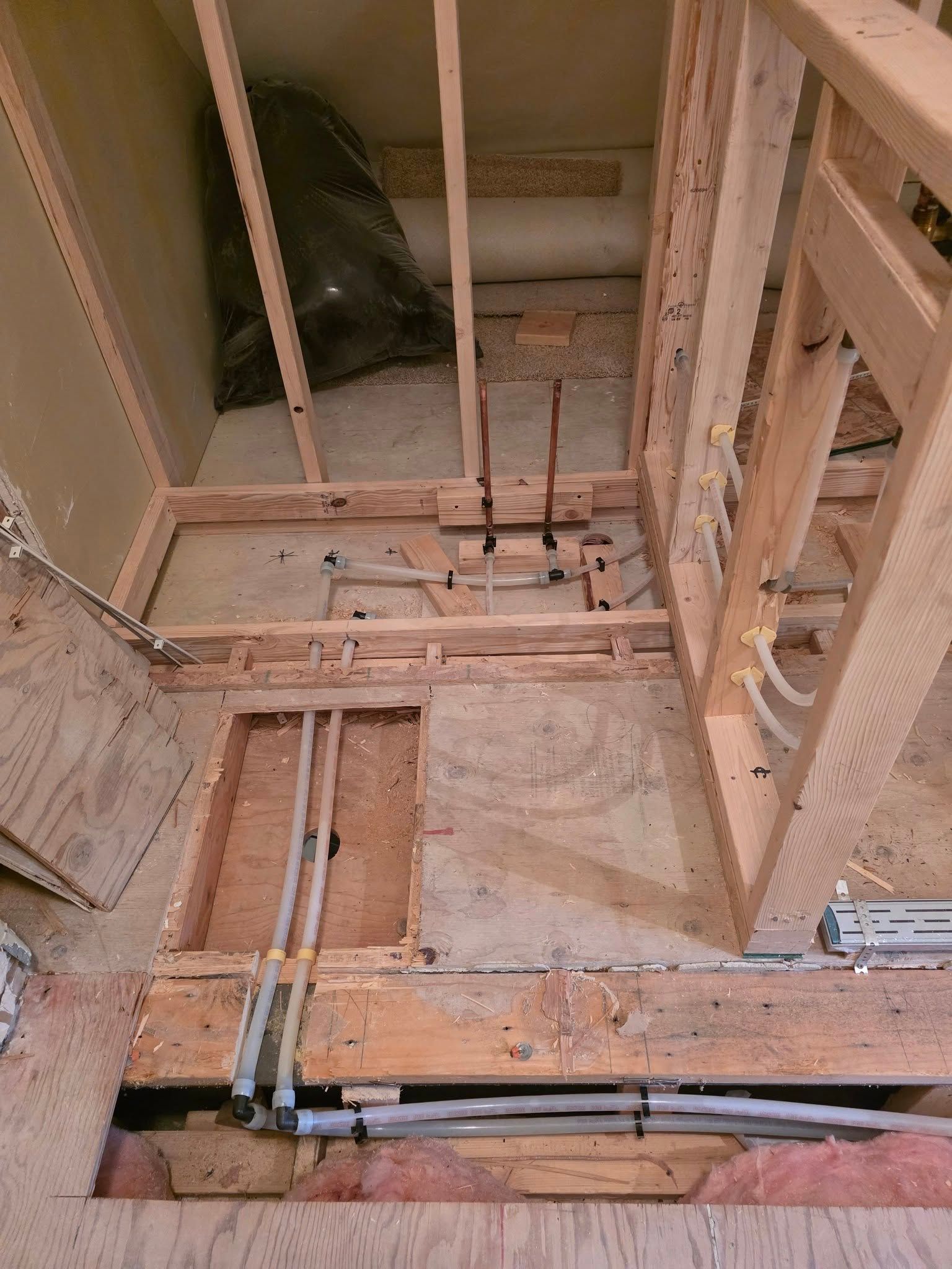 Interior view of a bathroom under construction with exposed wood framing, plumbing pipes, and insulation.