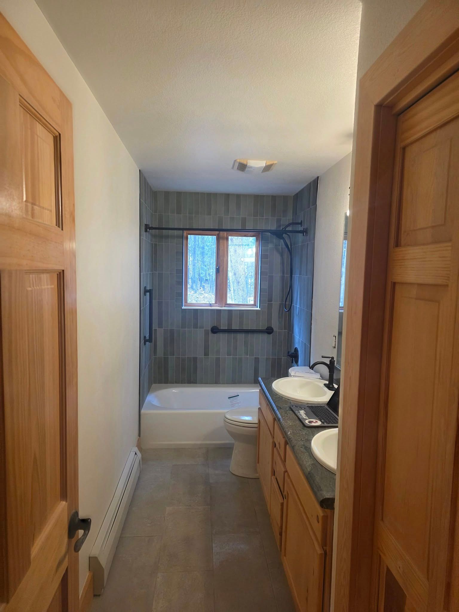 Bathroom with a tub, toilet, and vanity. Features gray tile, light wood cabinets, and two doors.
