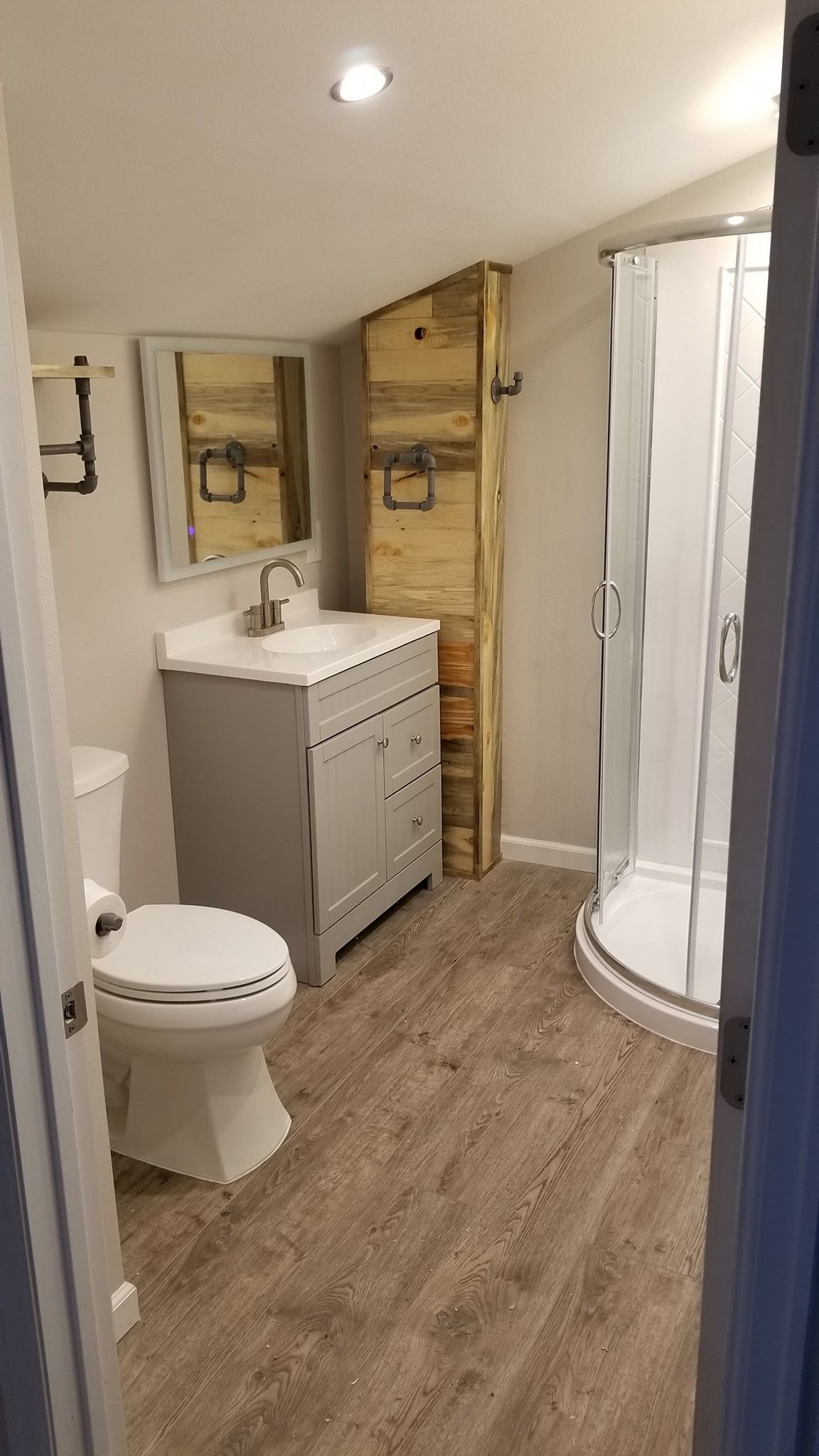 Bathroom with gray vanity, toilet, shower, wooden storage cabinet, and light wood-look flooring.