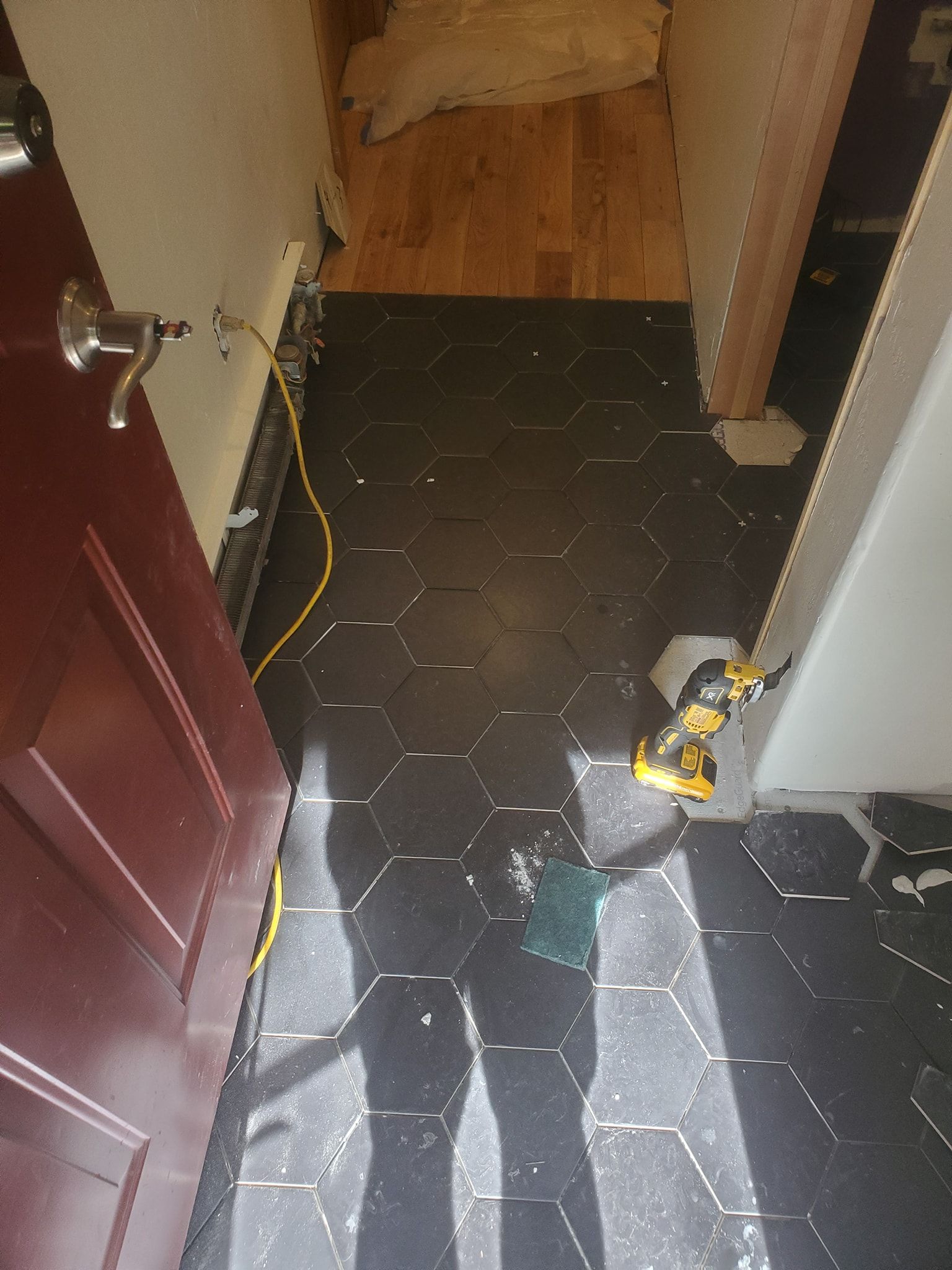 Black hexagon tile flooring installation in hallway. A drill and person's shadow visible. Door on the left.