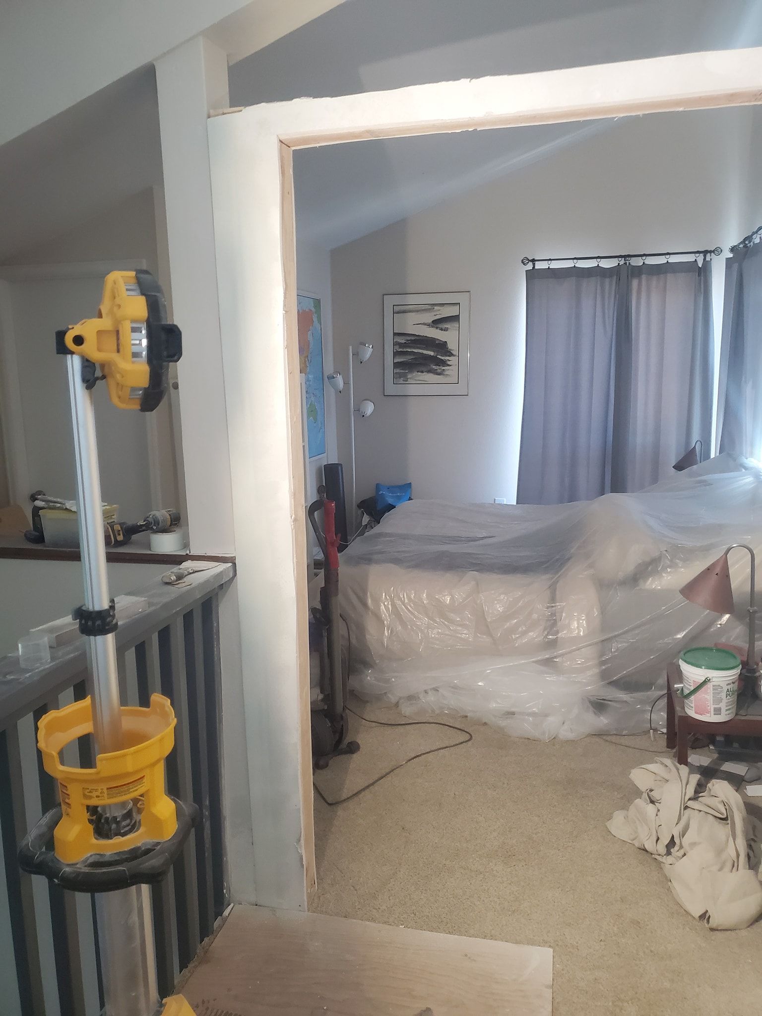 A doorway with fresh trim leads to a bedroom with bed covered in plastic, carpet, and curtains.