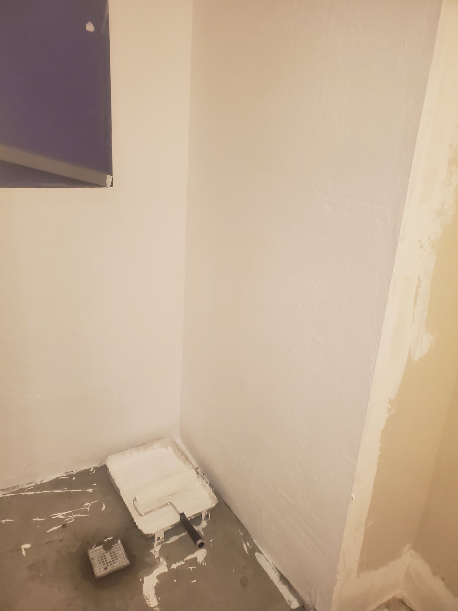 A corner of a room being painted white; paint roller and tray on the floor.