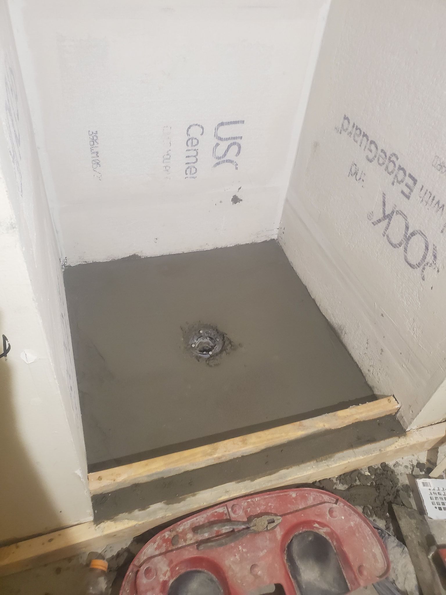 Shower construction in progress, gray cement base with drain, framed by wood and white wallboards.