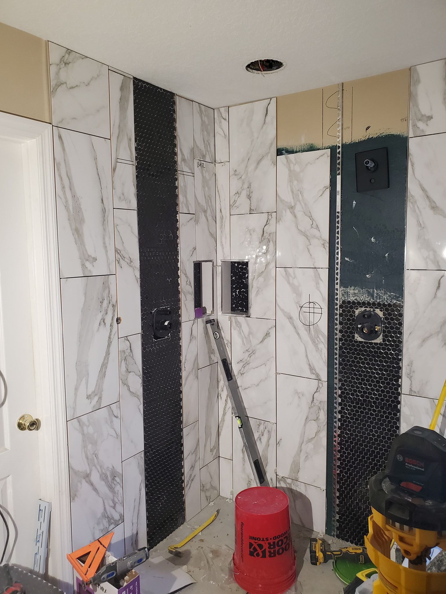 Bathroom remodel in progress: white marble tiles with black accent strip. Tools and bucket visible.