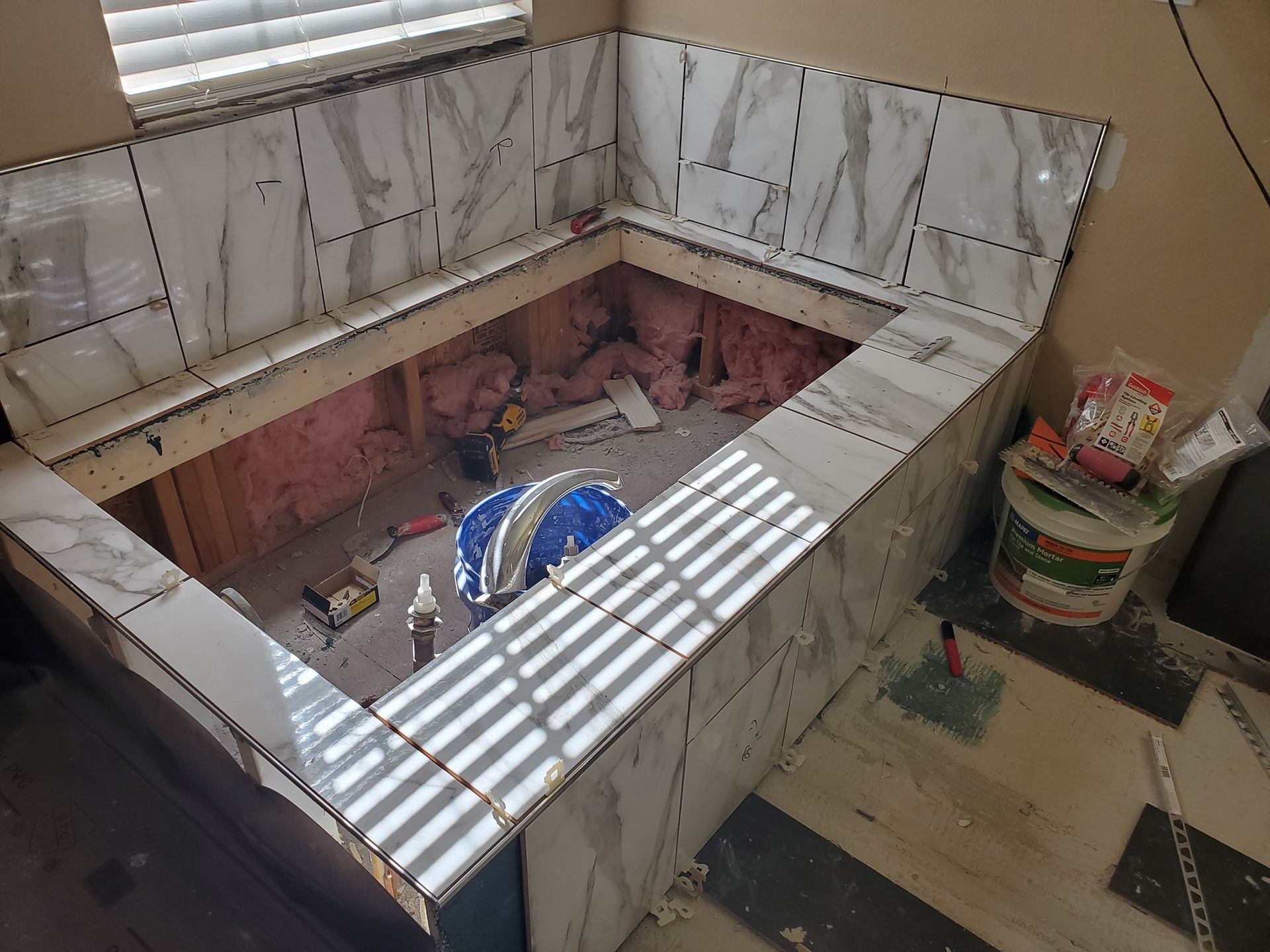 Bathroom tile installation in progress around a built-in tub. Marble-look tiles on walls and ledge. Pink insulation visible.