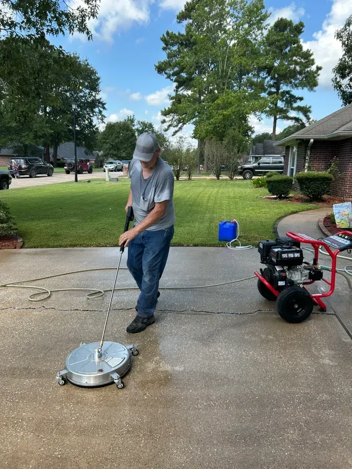 A person uses a circular surface cleaner attached to a pressure washer to clean a concrete driveway on a sunny day.
