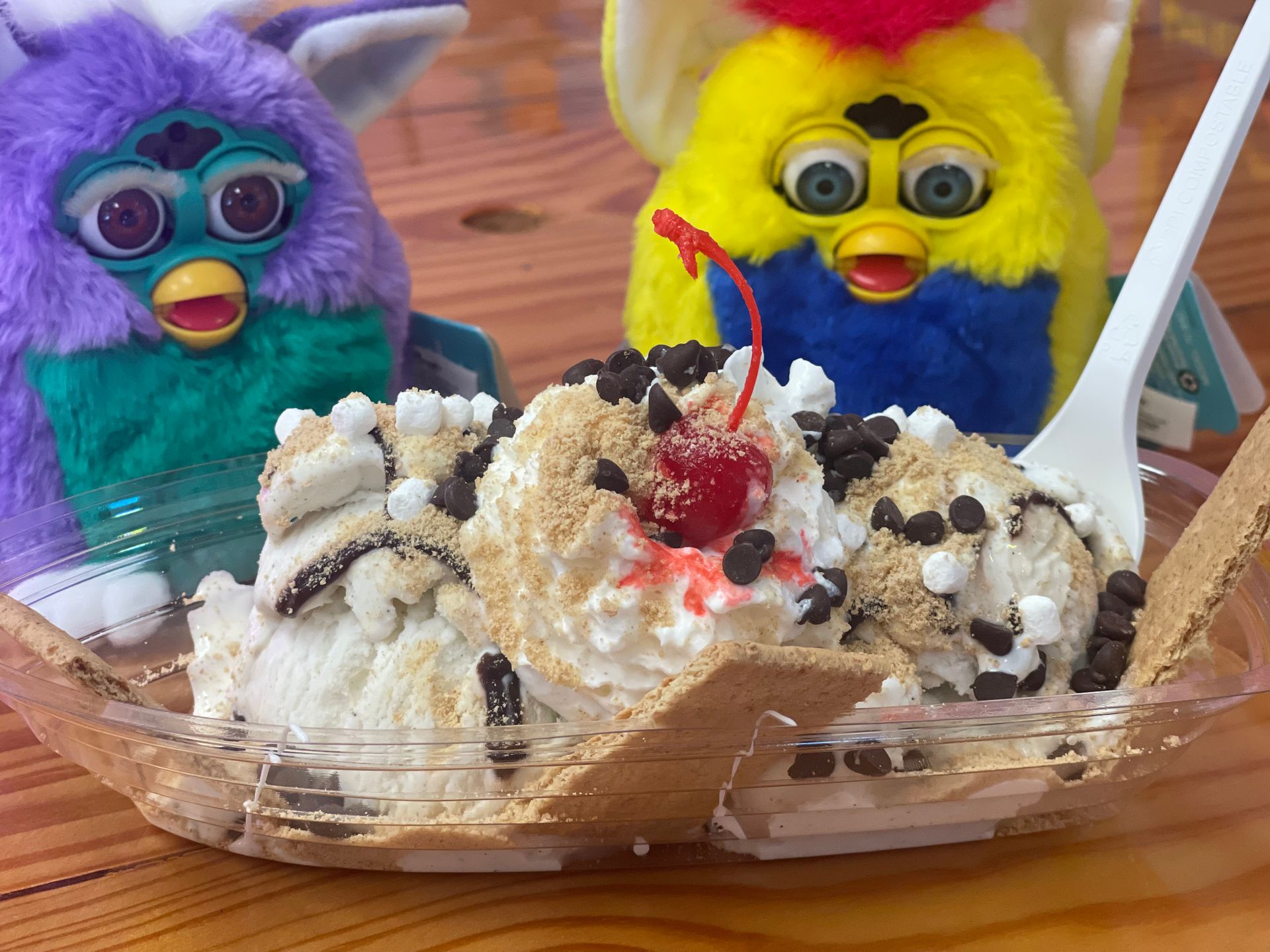 Two furby dolls are sitting next to a bowl of ice cream with a cherry on top.