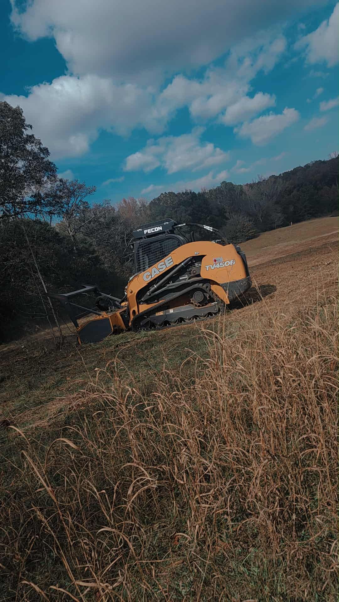 A yellow skid steer tractor on a grassy hillside under a blue sky.