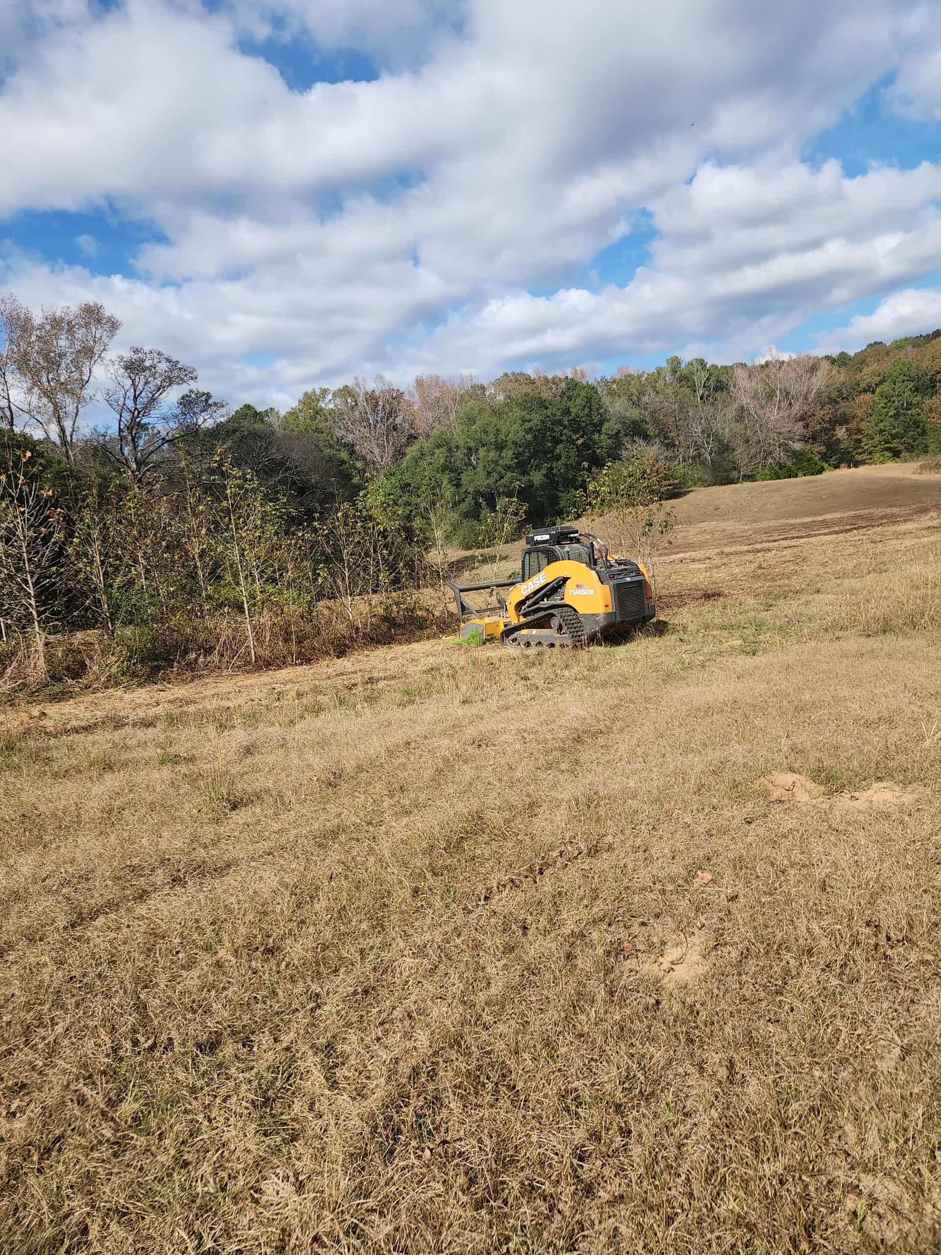 Small, yellow tracked machine on a grassy hill; trees and a partly cloudy sky are in the background.