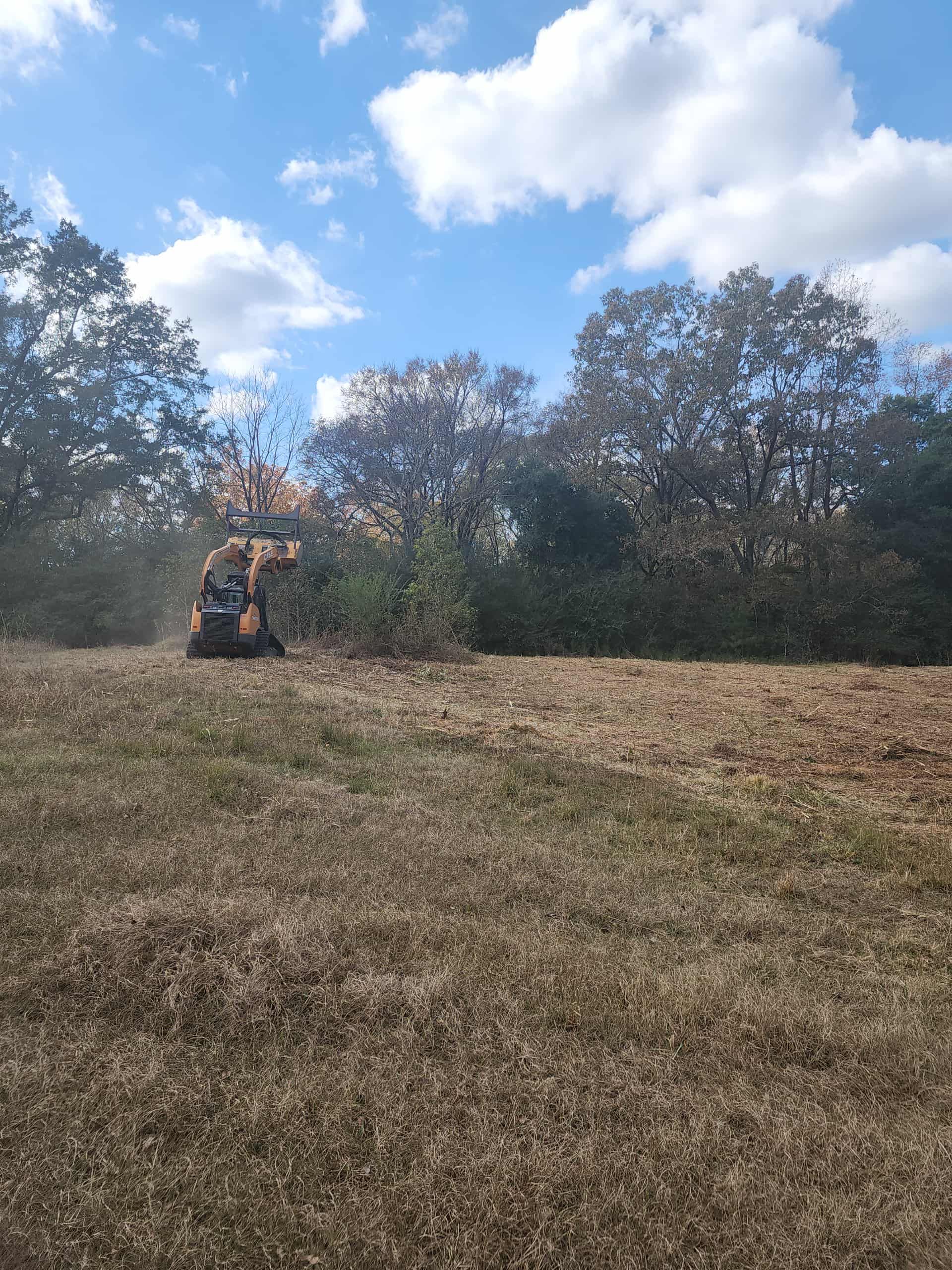 A small orange machine clearing brush in a field. Trees line the background under a blue sky with clouds.