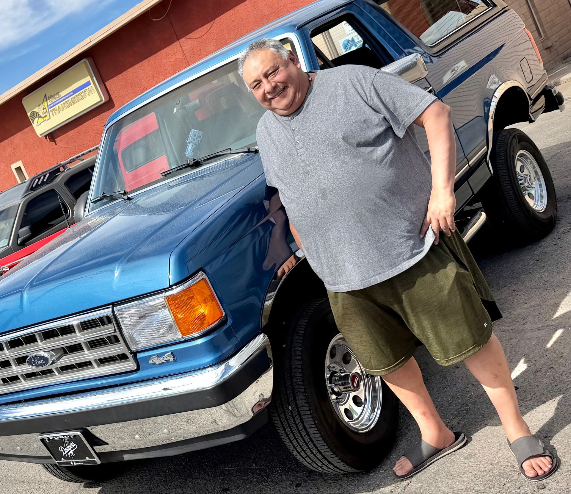 Man smiles next to a blue Ford Bronco. He wears a gray shirt, green shorts, and sandals.