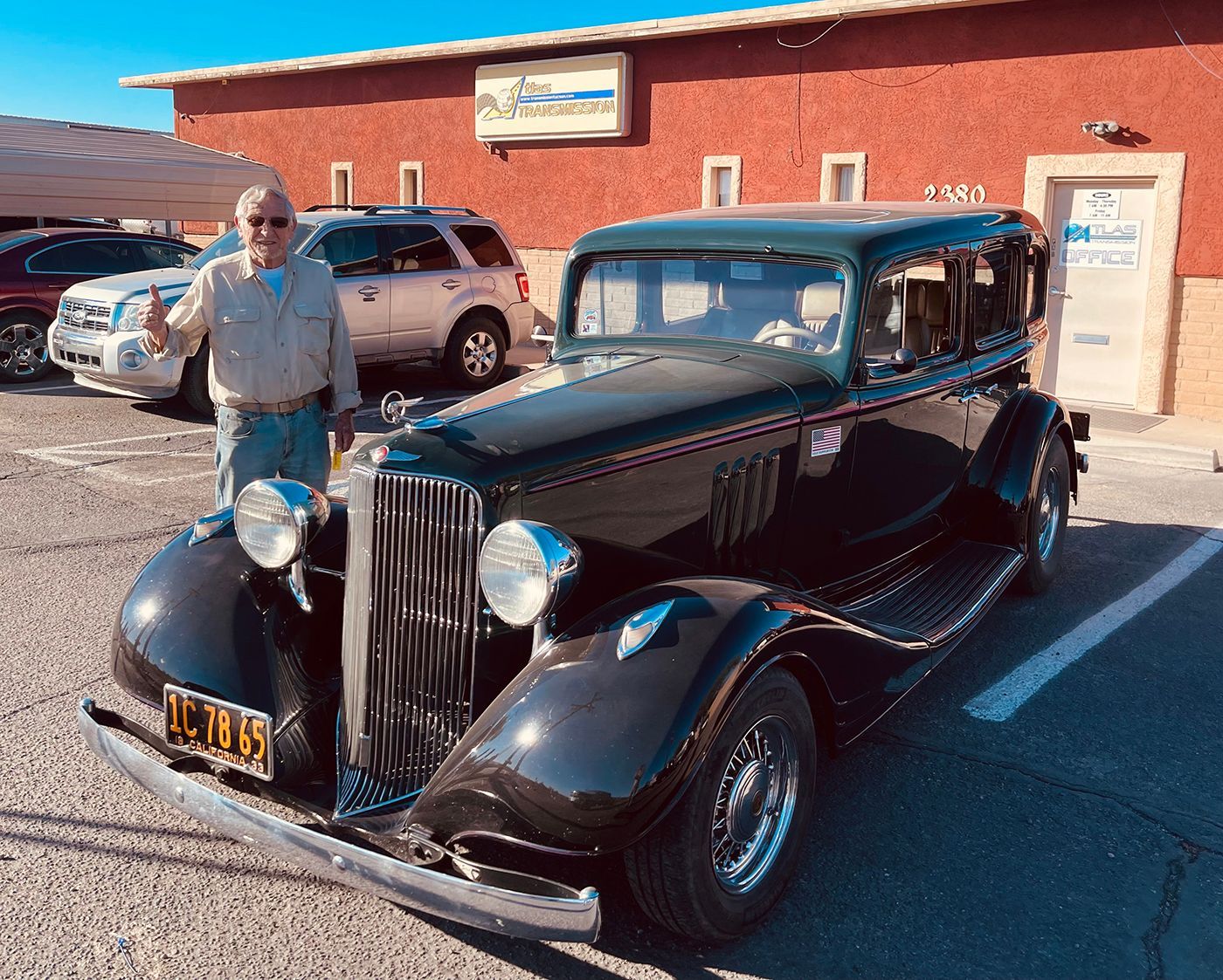 Man stands next to a black vintage car. Building in background, parked cars, sunny day.