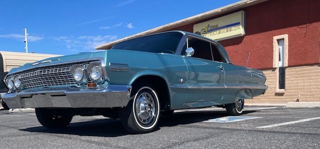 A turquoise 1963 Chevrolet Impala parked in front of a brick building on a sunny day.