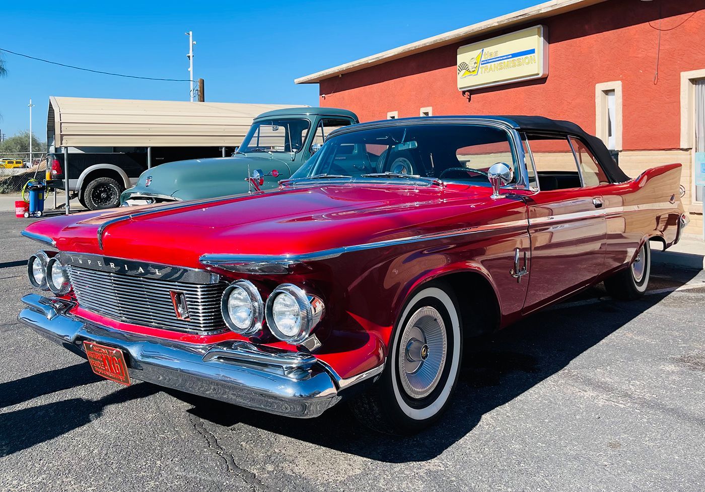 Red vintage convertible car parked in front of a building; another truck is behind.