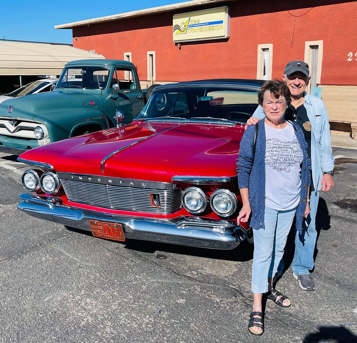 Couple poses with a red classic car, green truck in background, outside a red building.