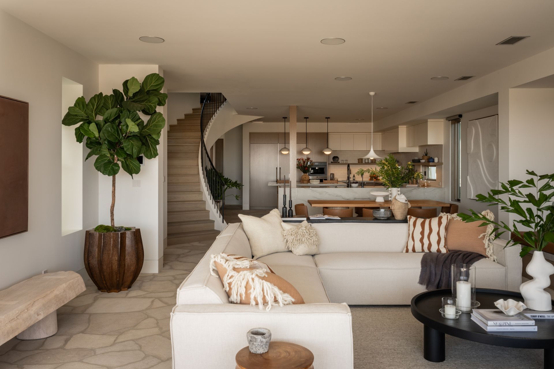 A bright living room with a cream sectional sofa, a large potted fiddle leaf fig, and stairs leading to an open kitchen.