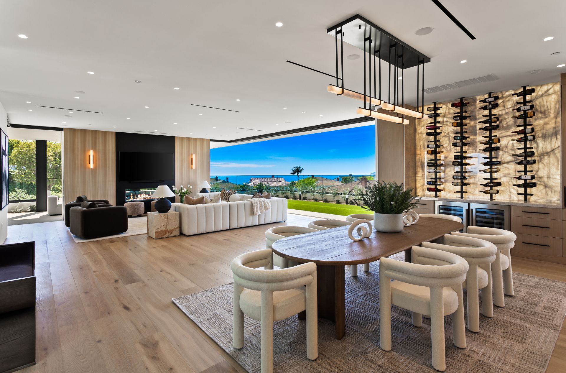 A modern, open-plan living and dining room with a large wooden table, cream chairs, a wine wall, and an ocean view.