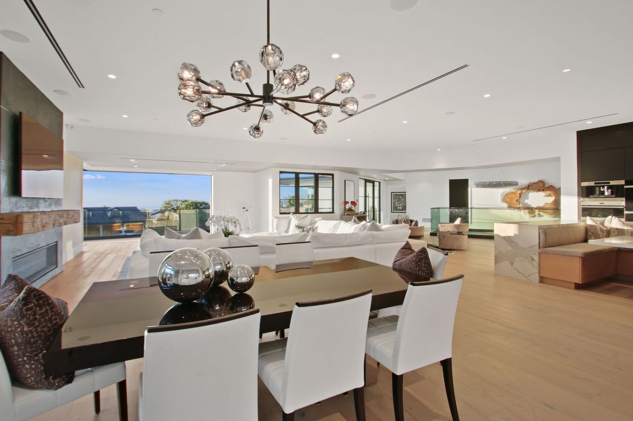 A bright, modern open-concept living and dining area with a glass-topped dining table, white chairs, and a chandelier.