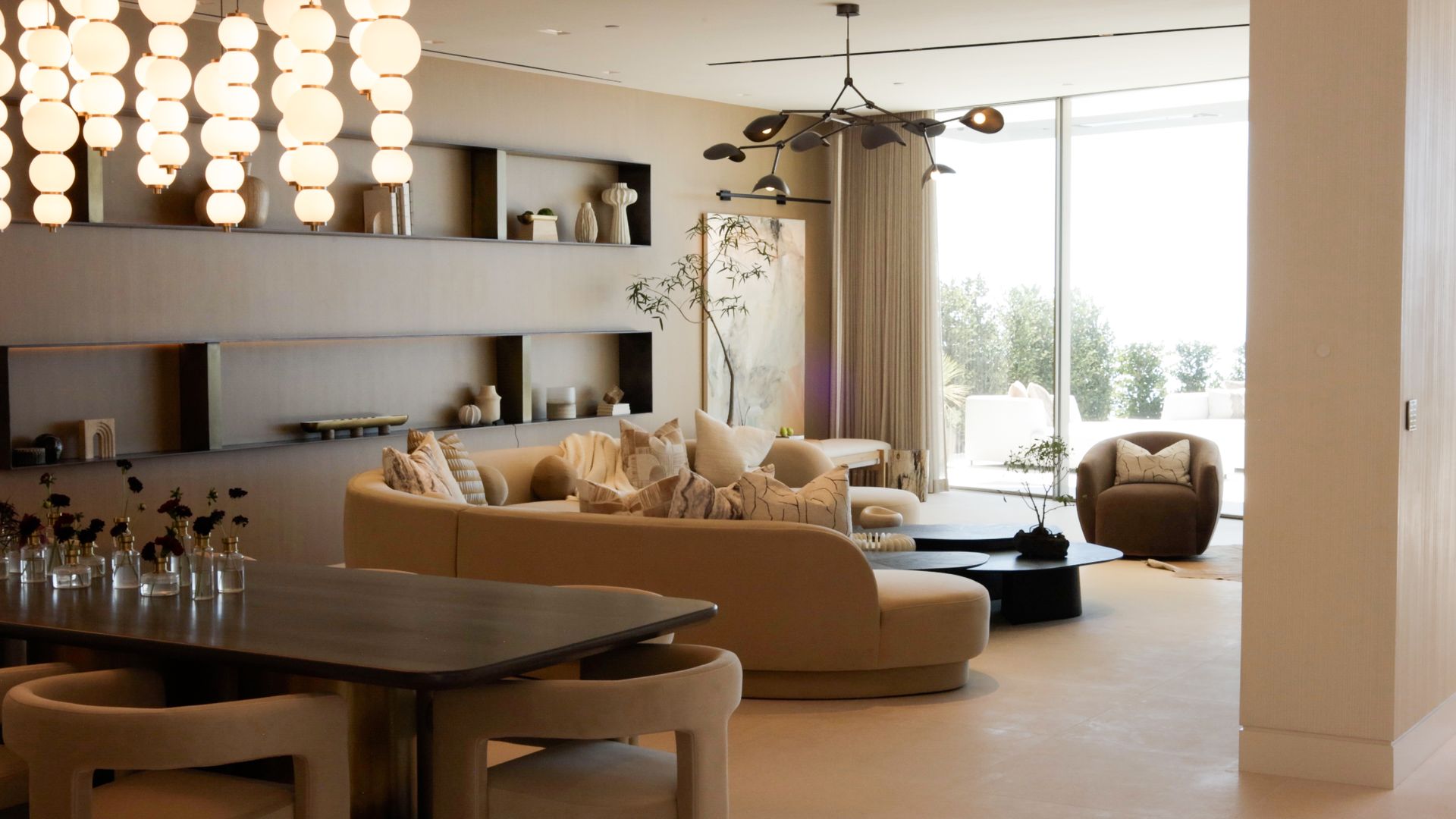 A modern living and dining room with a neutral palette, curved sofa, dark wood dining table, and contemporary lighting.