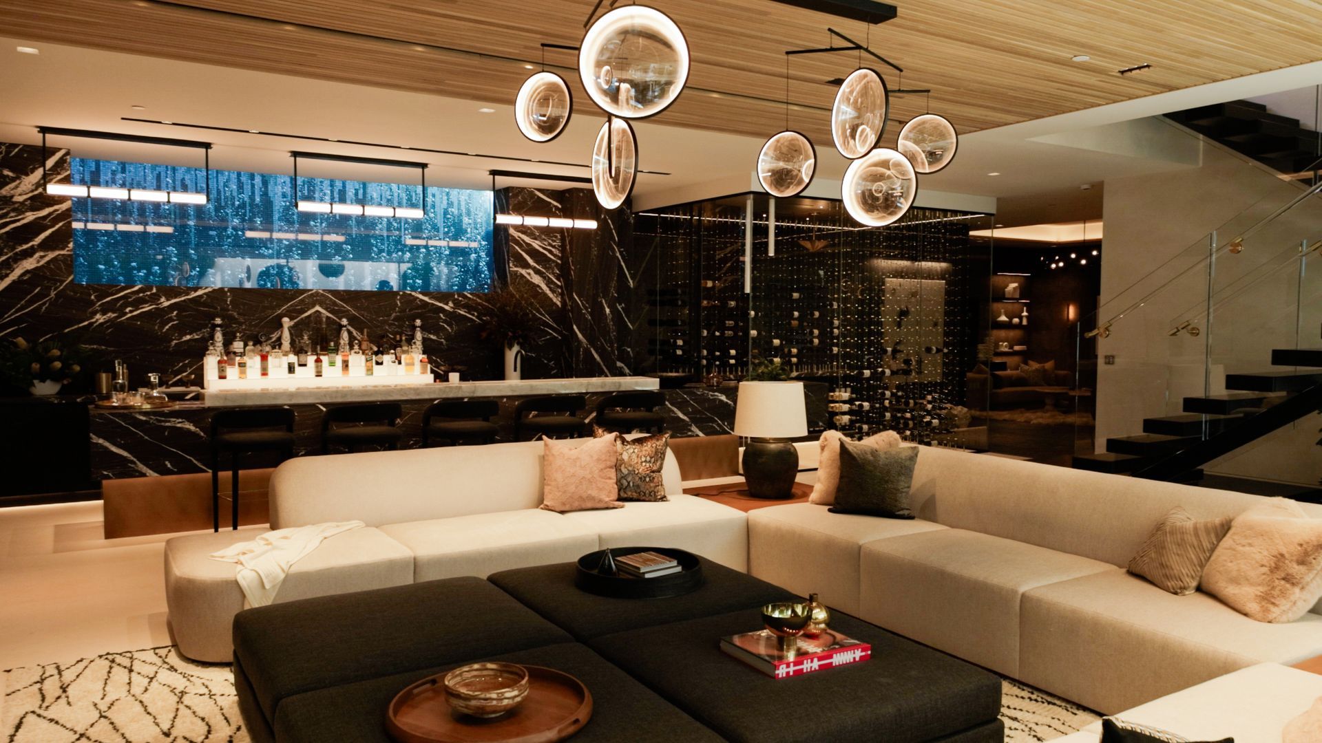 A modern, open-plan living room with a large beige sectional, black ottoman, dark marble walls, and unique globe lighting.