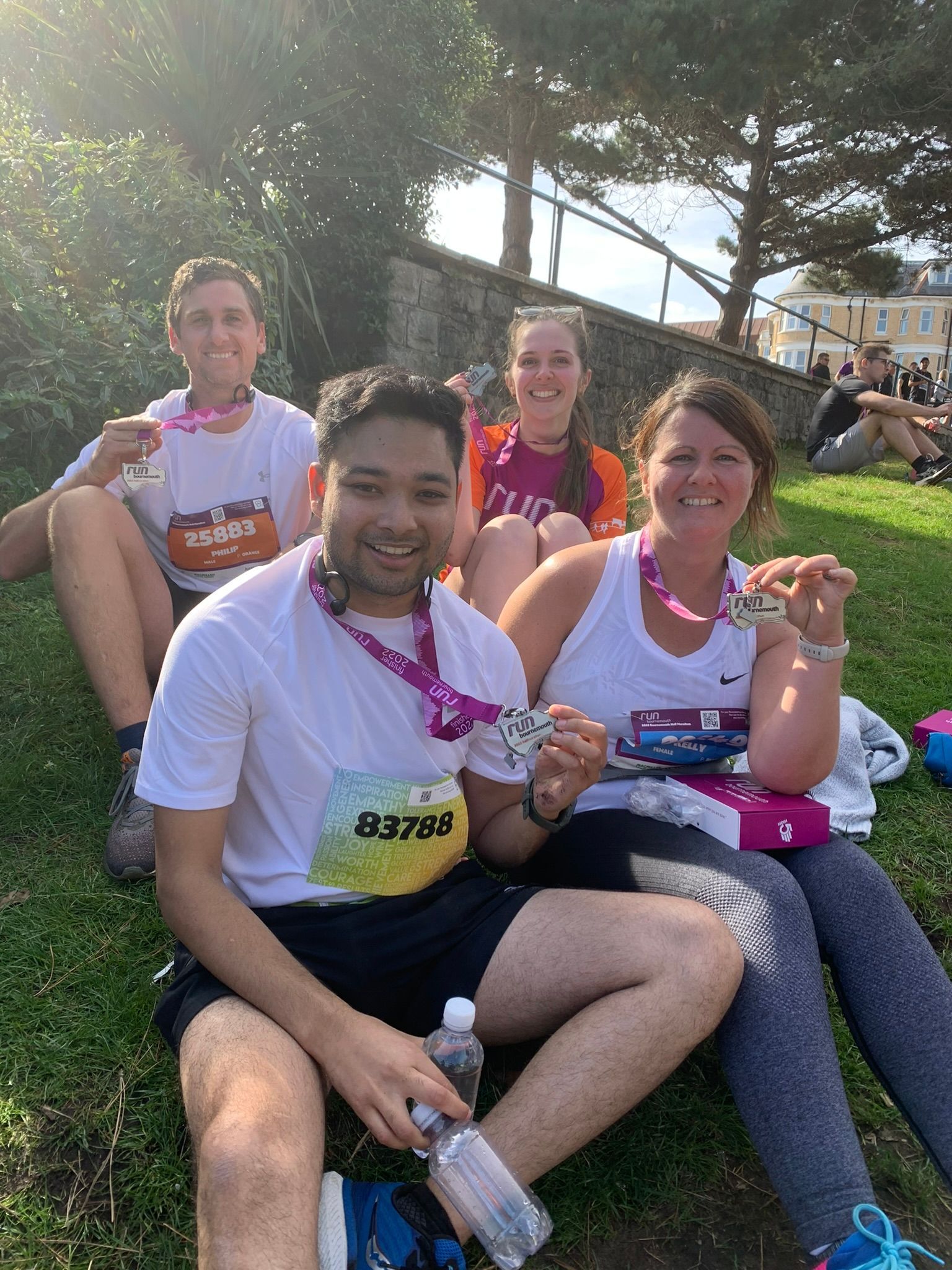 Four people sitting on grass, smiling and holding medals, after a race.