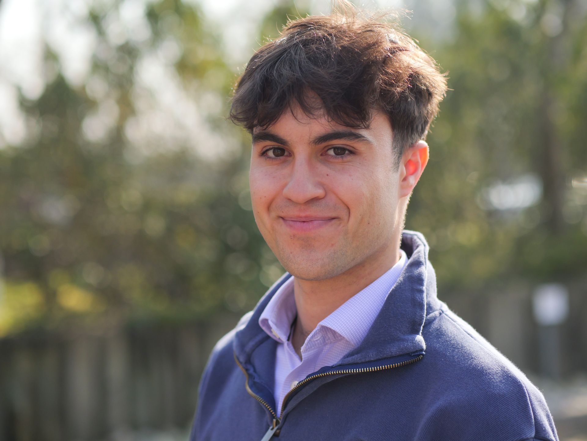 A person with dark, wavy hair, wearing a light purple shirt and blue sweater, smiles in an outdoor, wooded setting.