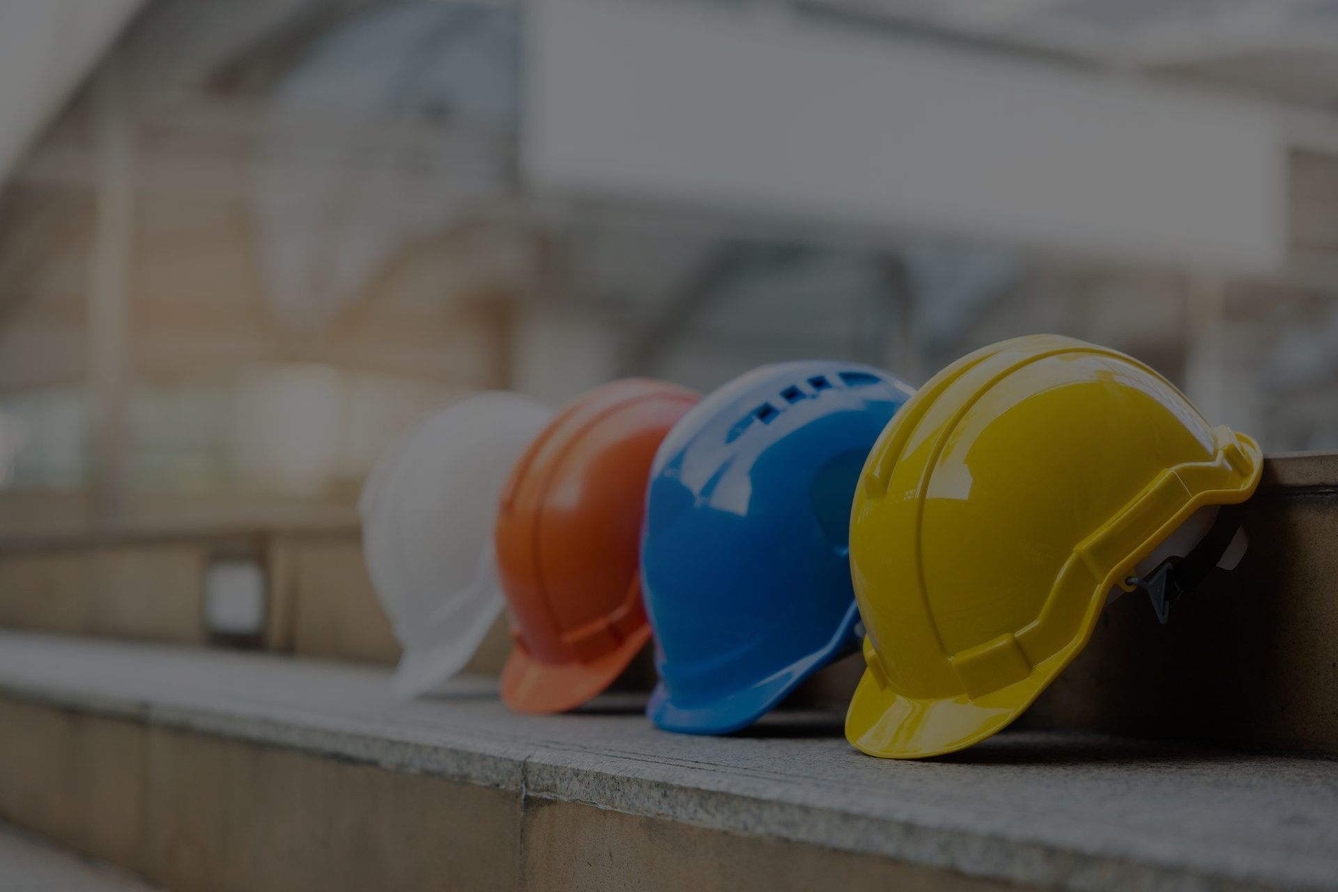 Four colorful construction hard hats (white, orange, blue, and yellow) lined up on a ledge, possibly outdoors.