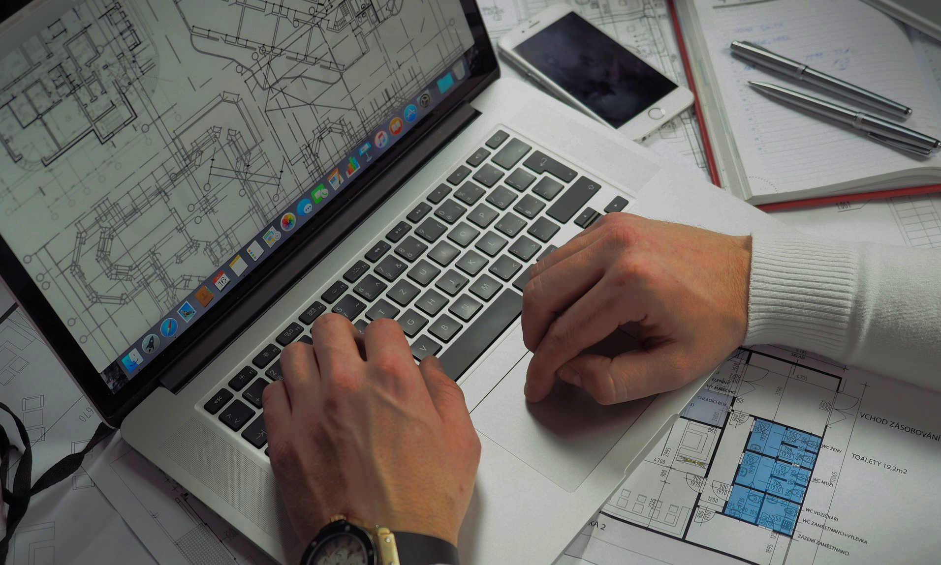 Hands typing on a laptop, architectural blueprints, phone, pens, and watch on a desk.