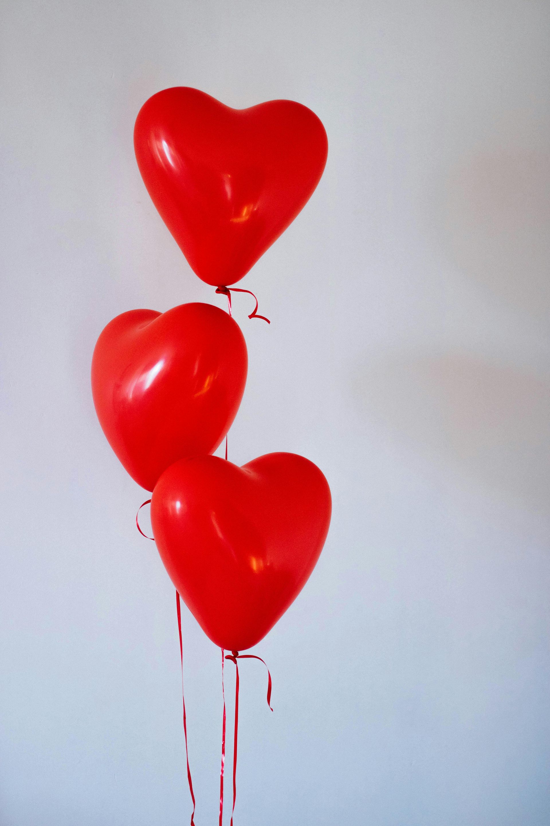 Three red heart-shaped balloons floating against a plain white background, red ribbons dangling.