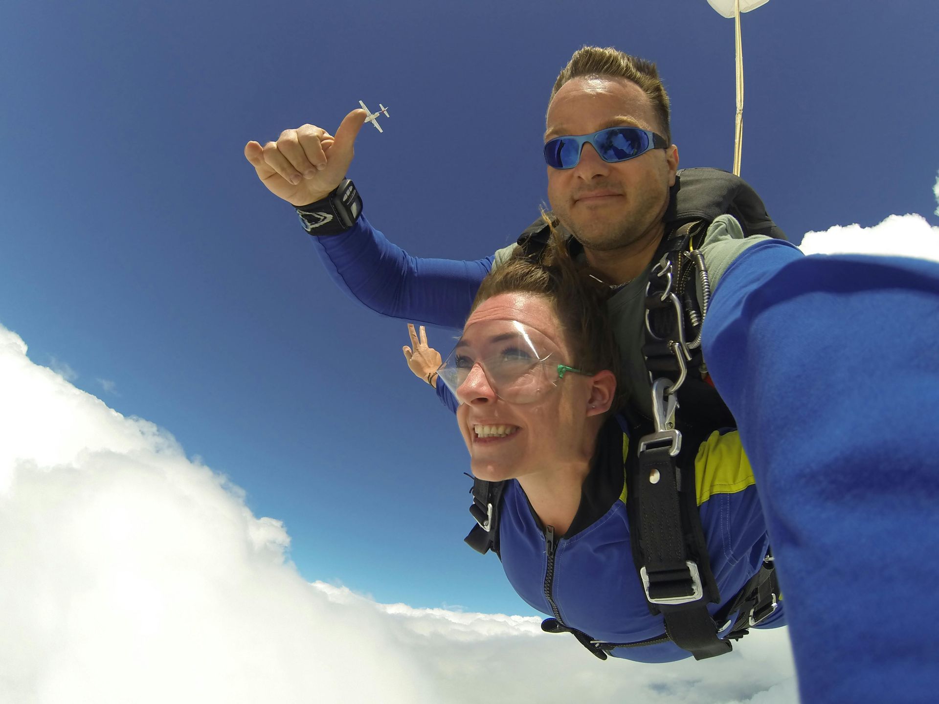 Tandem skydivers in blue jumpsuits, smiling, flying above clouds with a blue sky. One gives a thumbs up.