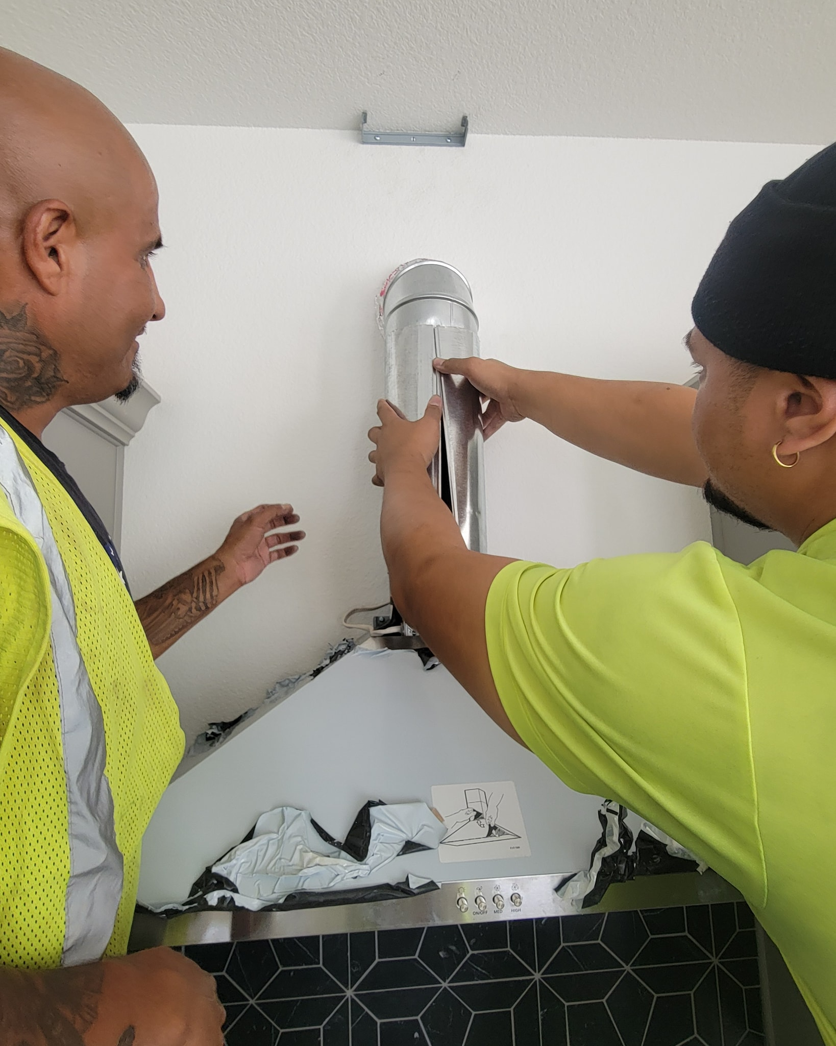 Two people installing a metal vent pipe to a kitchen range hood on a white wall.