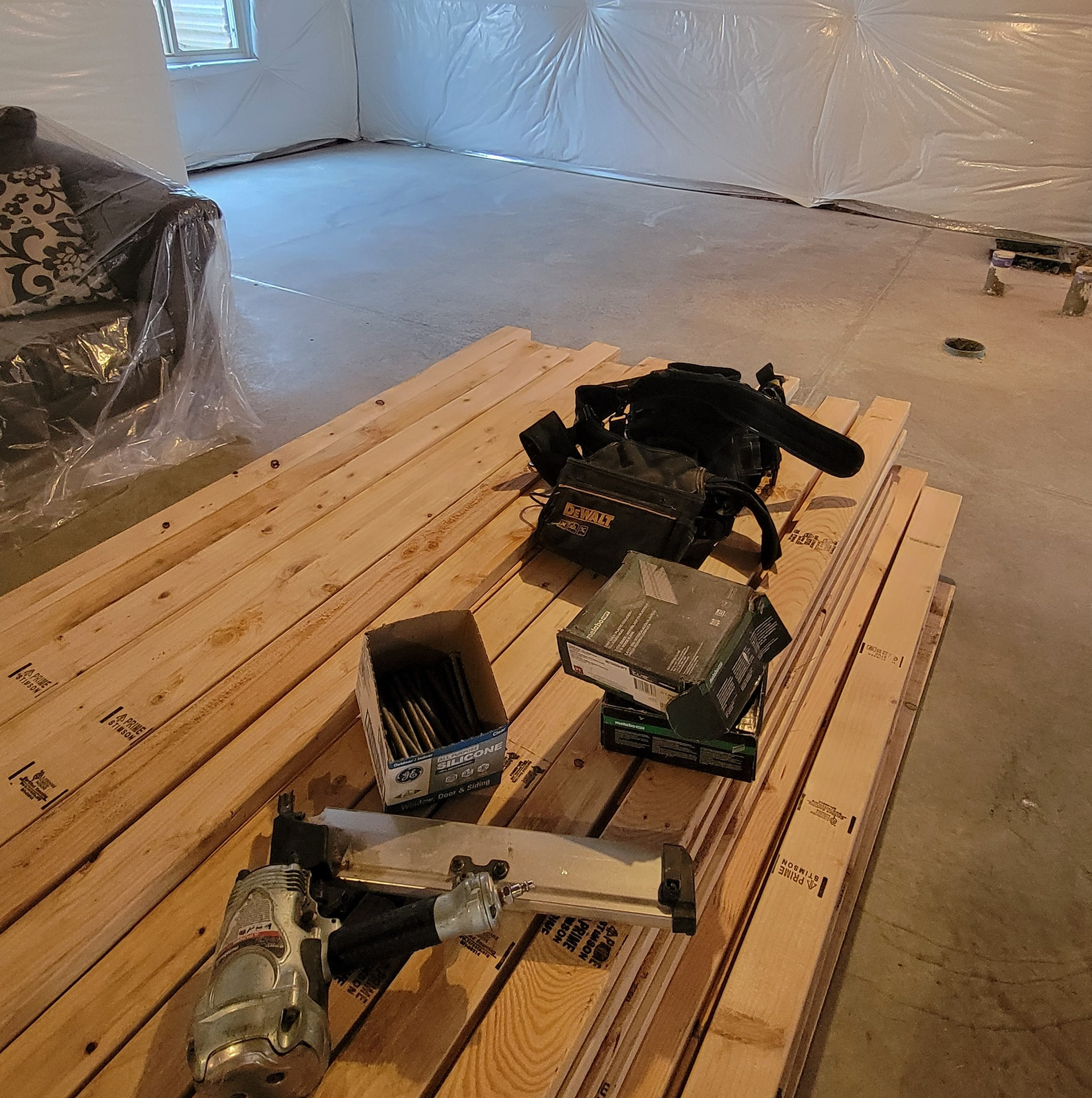 Basement renovation: wood planks, nail gun, tool bag, and boxes on concrete floor.
