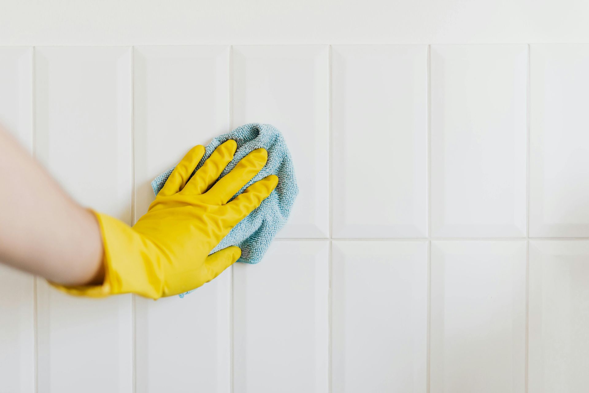 Hand in yellow glove wiping white tiled wall with blue cloth.
