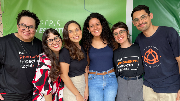 Seis pessoas estão de pé, sorrindo juntas, em frente a uma parede verde onde se exibem as palavras