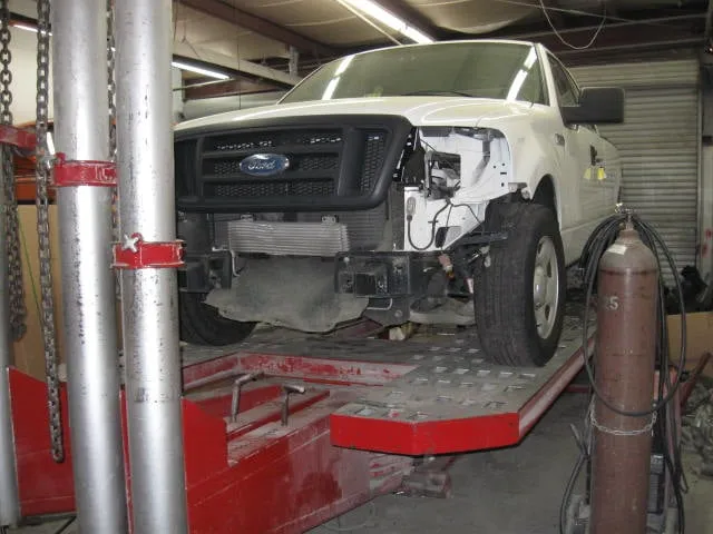 White Ford pickup truck on a car lift inside a repair shop, front-end damage visible.