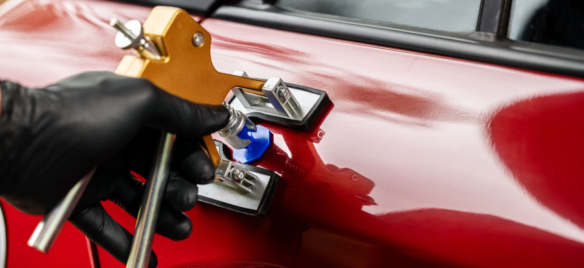 Hand using a dent puller tool on a red car door to remove a dent.