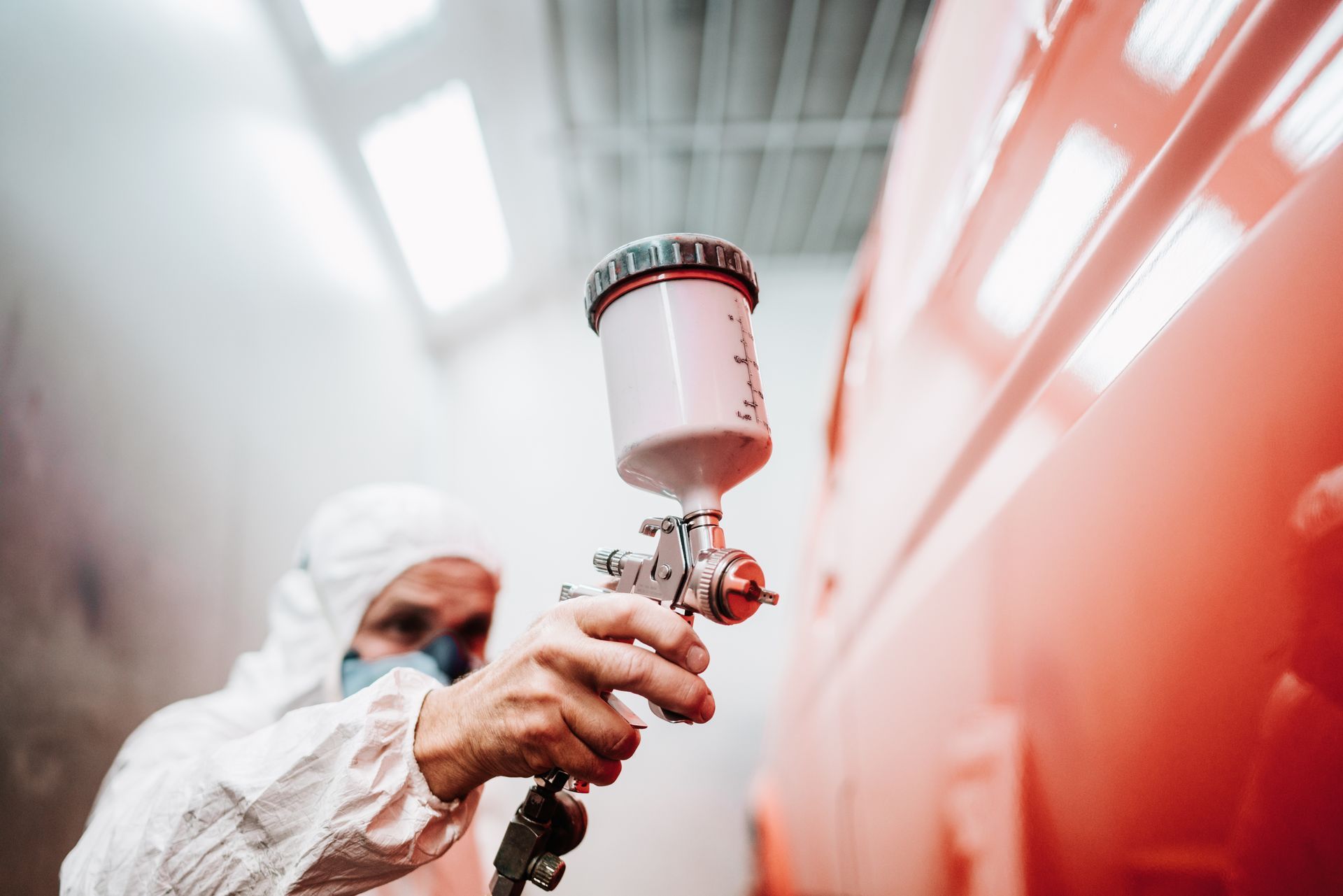 Person in protective suit sprays red paint onto a vehicle in a spray booth.