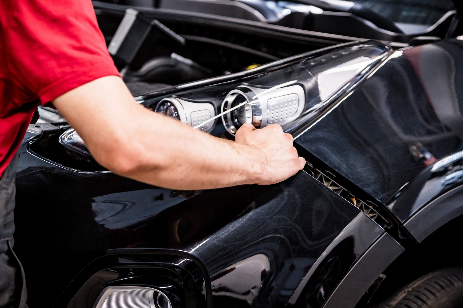 Mechanic in red shirt inspecting a black car's hood, likely in a garage.