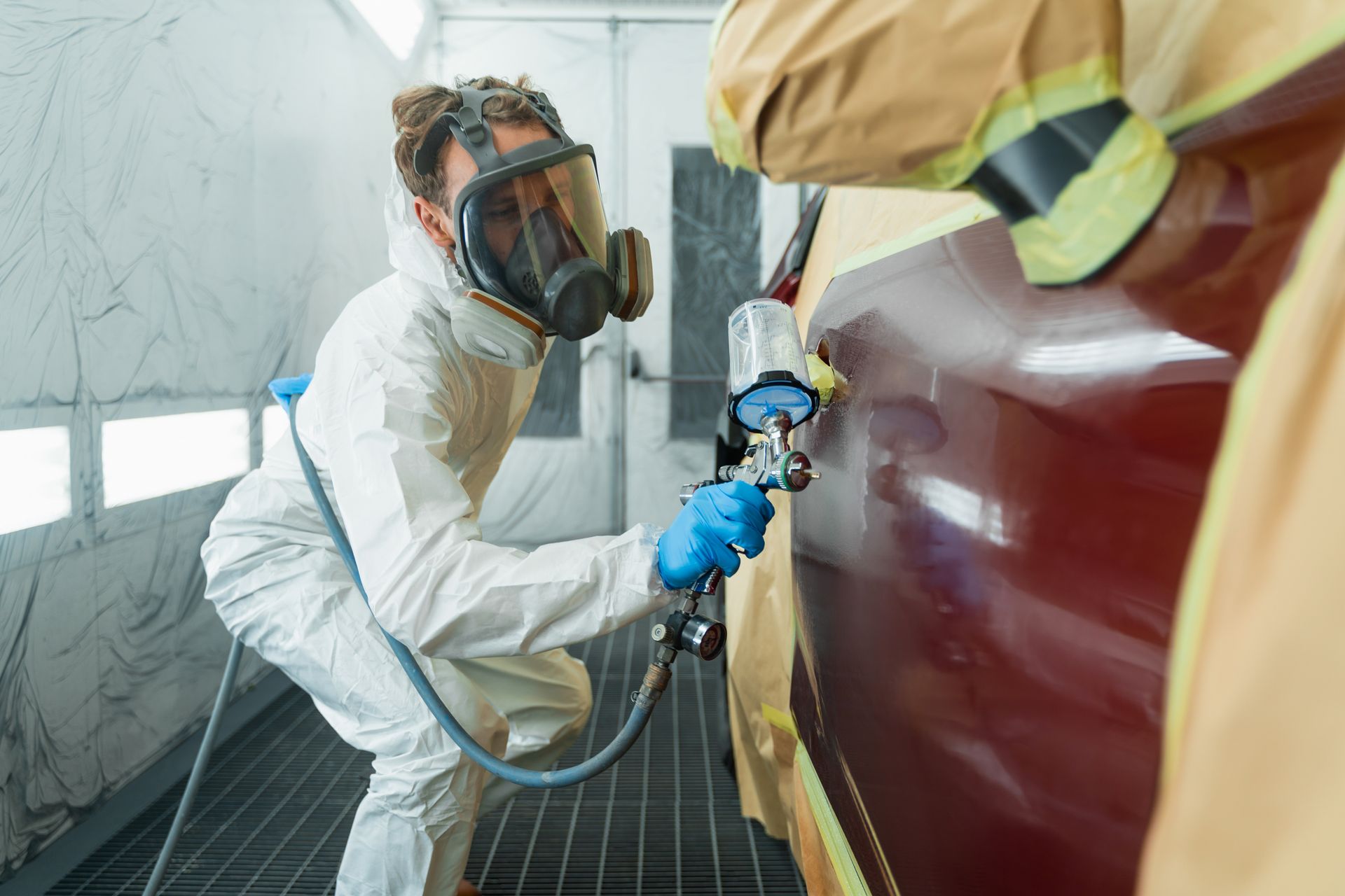 Person in protective suit and respirator sprays paint on a car in a paint booth.