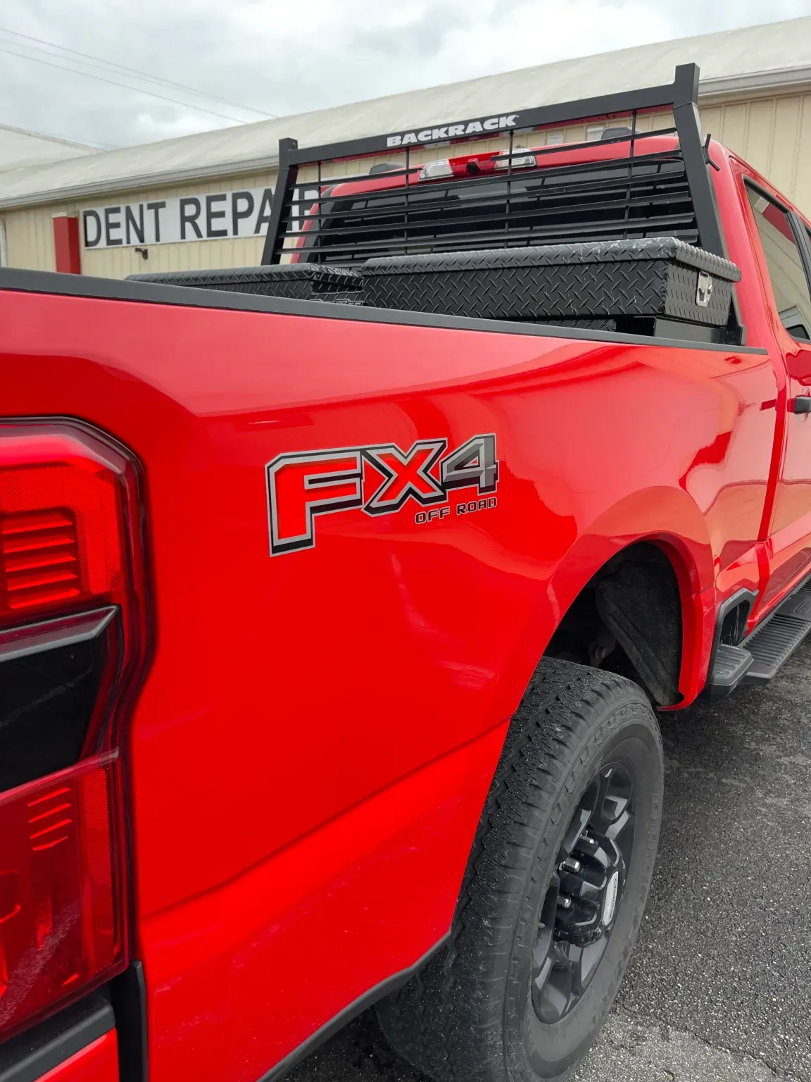 Red Ford truck with FX4 logo, black toolbox and rack, parked outside a dent repair shop.