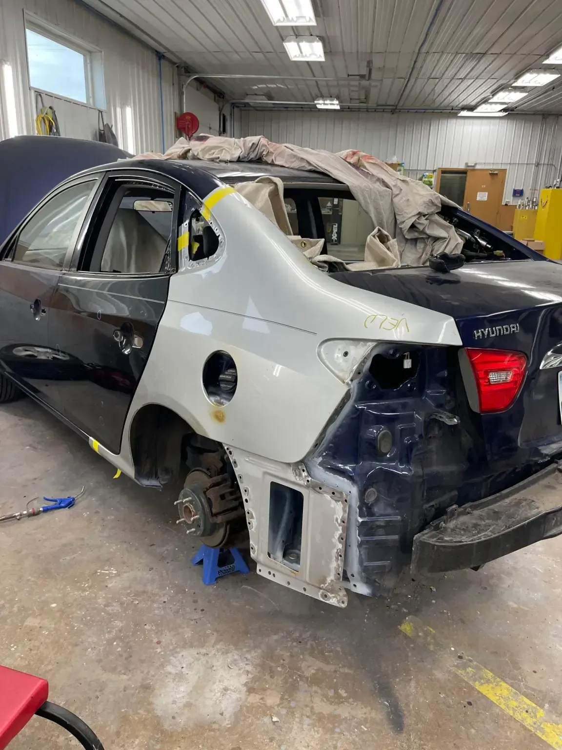 A car with a damaged rear panel being repaired in a garage; dark blue and silver panels.