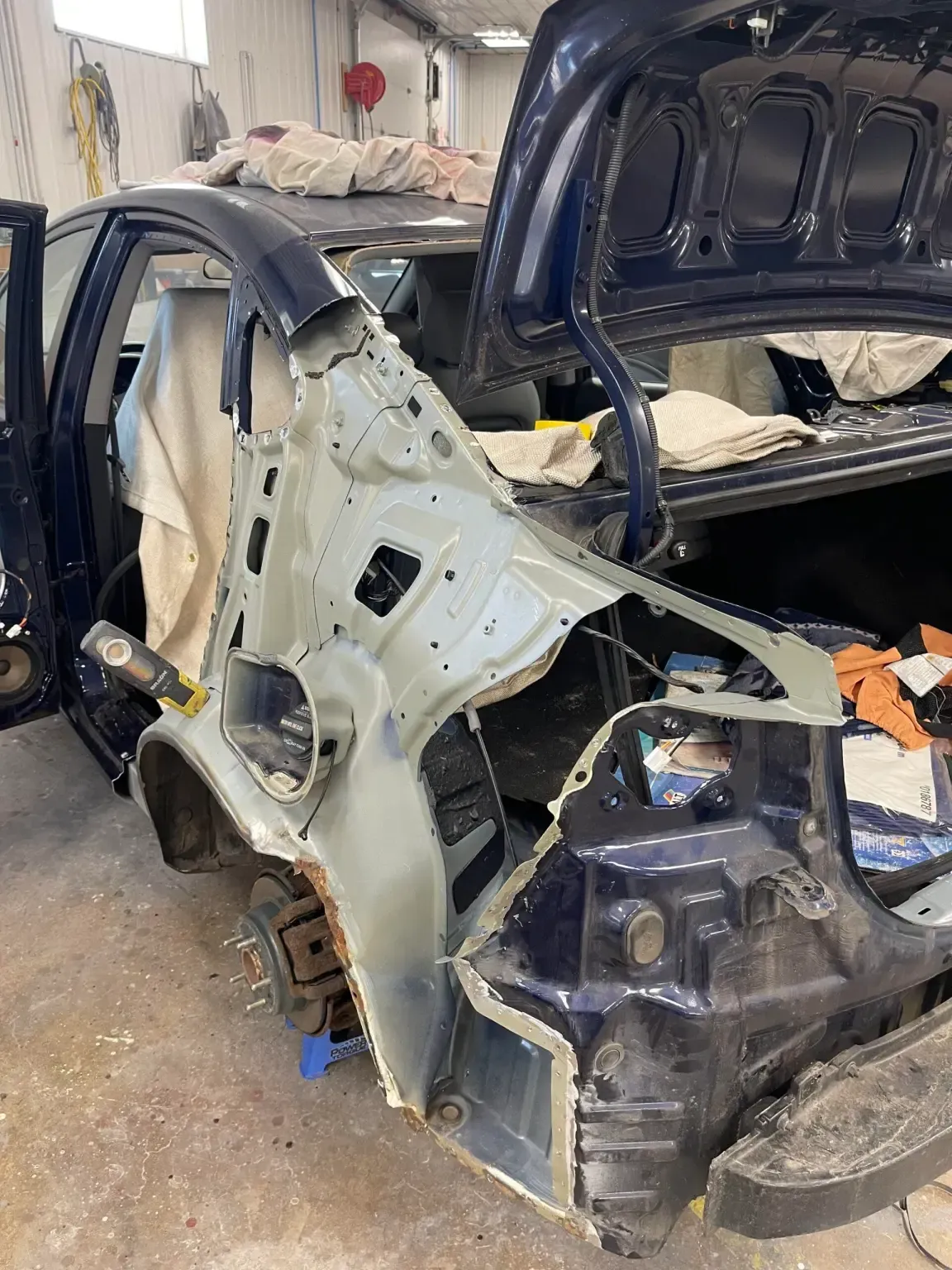 Car body repair: blue car with the rear quarter panel removed, revealing internal structure. Garage setting.