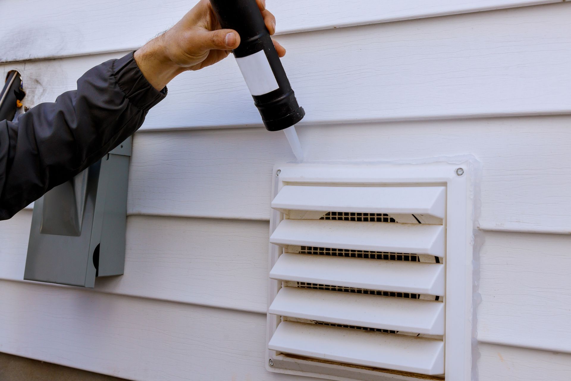 Person sealing a white vent on white siding with a caulk gun.