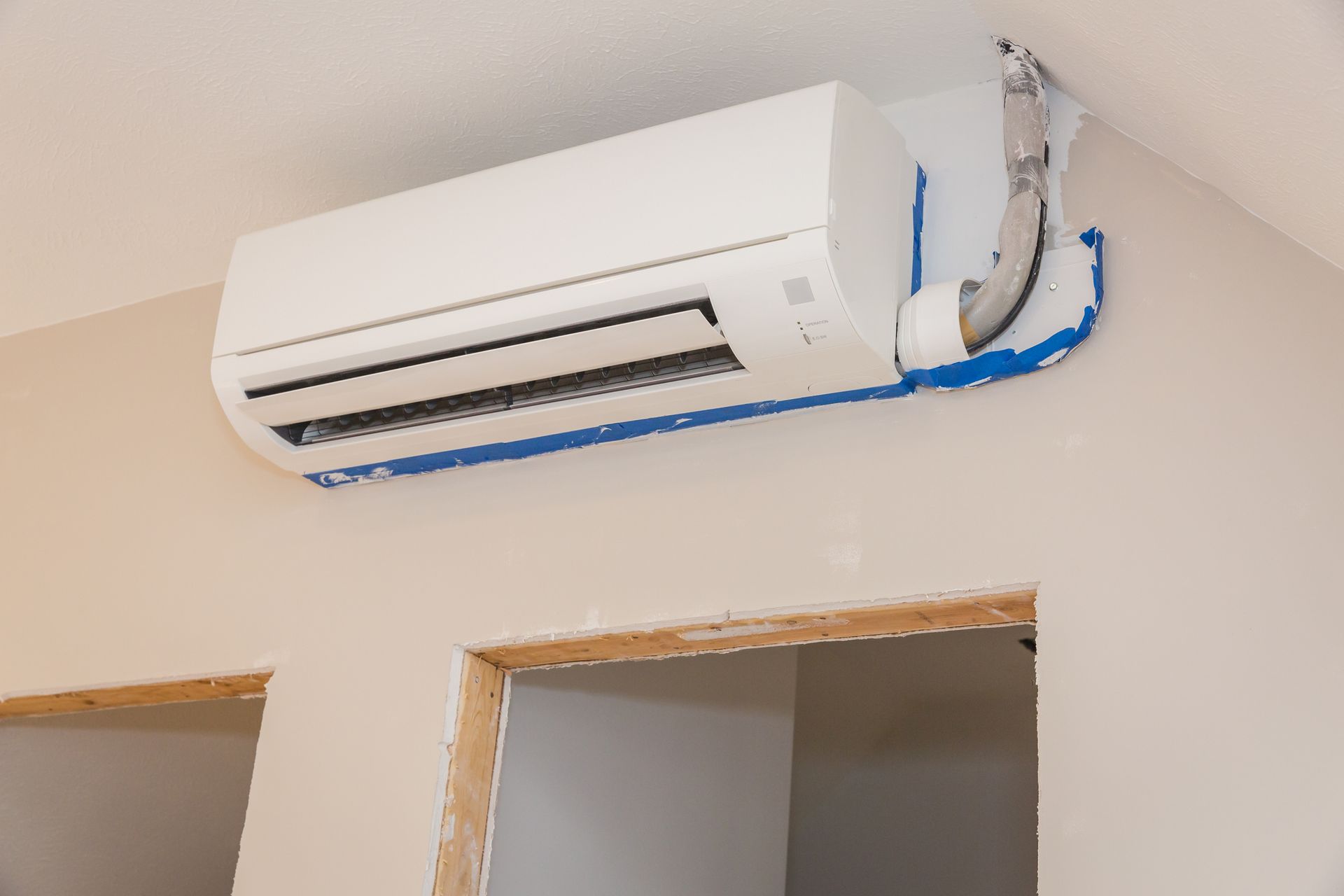 White air conditioner unit mounted on a light-colored wall, with blue tape covering pipes near an opening.