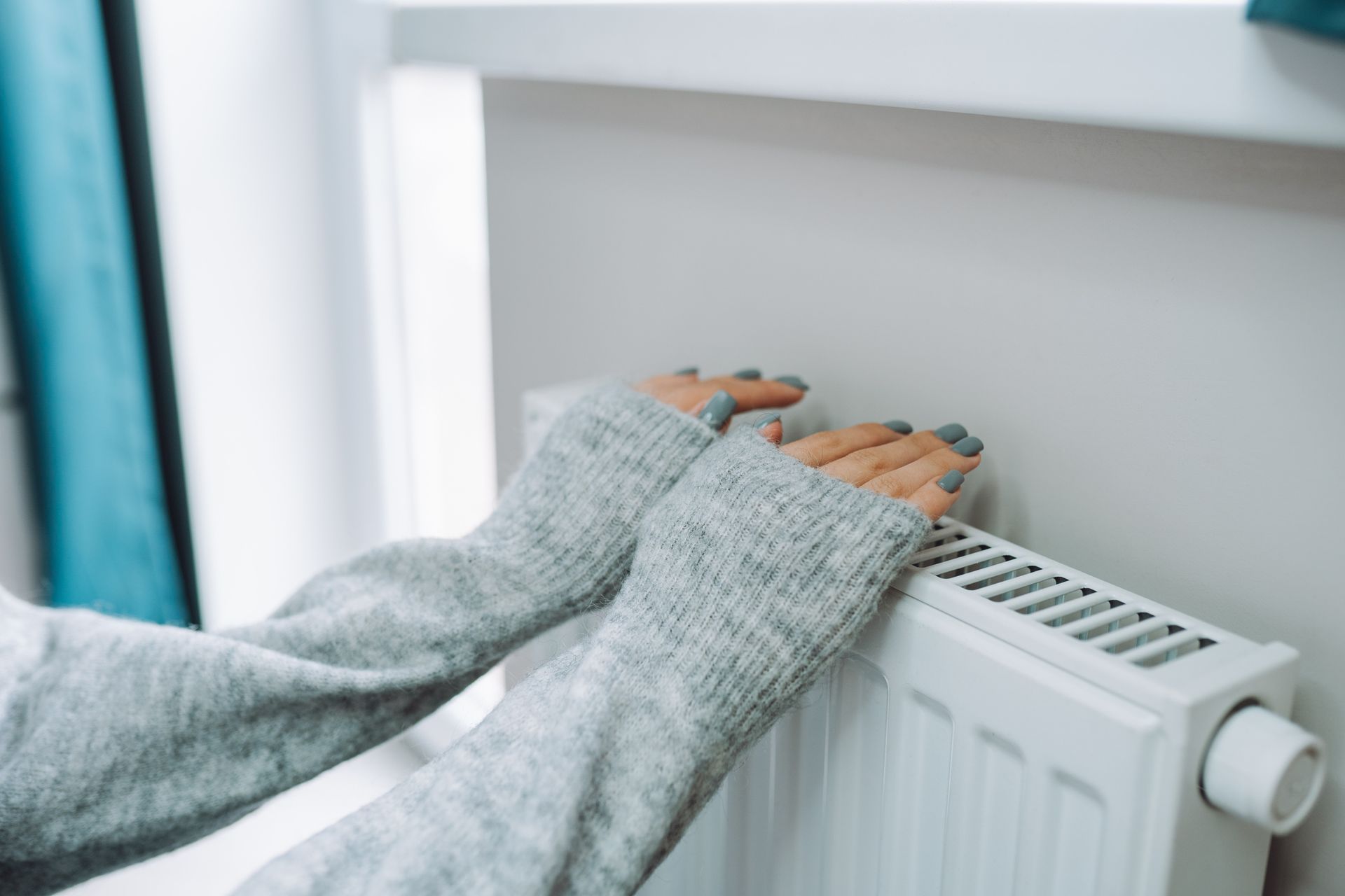 Hands warming over a white radiator near a window, wearing a gray sweater.